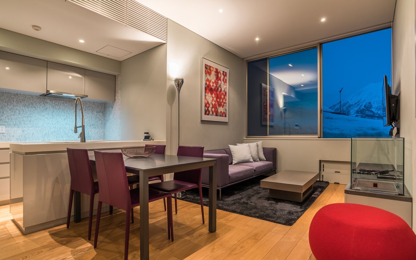 Une cuisine moderne dotée d’un îlot central intégré, un coin repas avec une table et des chaises, un canapé et une grande fenêtre offrant une vue sur un paysage enneigé.