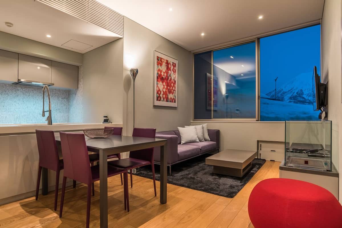 A modern kitchen with a built-in island, a dining area with a table and chairs, a sofa, and a large window with a view of a snowy landscape.