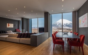 A modern interior with a sofa, dining table, chairs, and large windows offering a view of a snow-capped mountain.