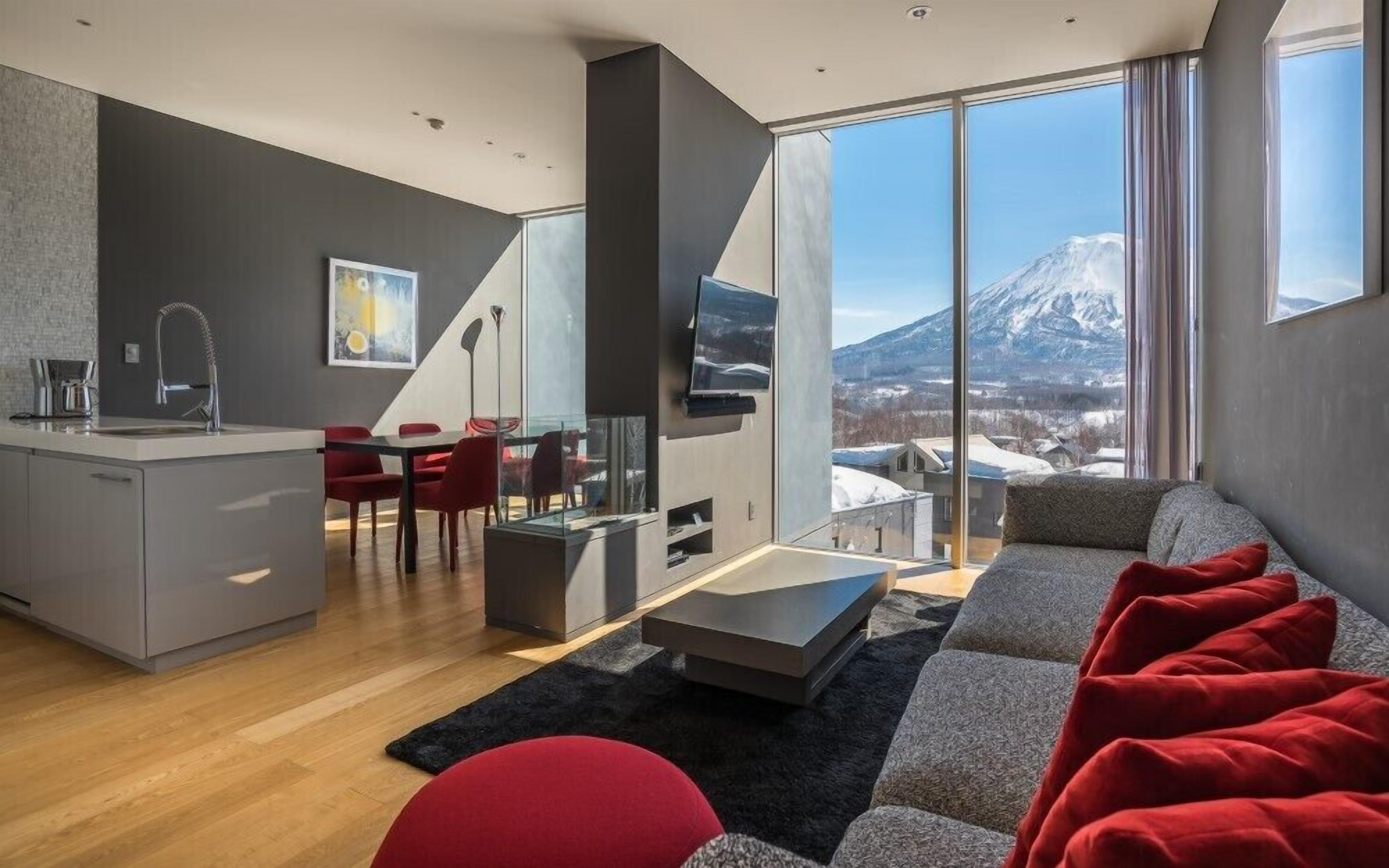 Un salon moderne comprenant un canapé, des fauteuils rouges, une table à manger, un coin cuisine et une grande fenêtre donnant sur les montagnes.