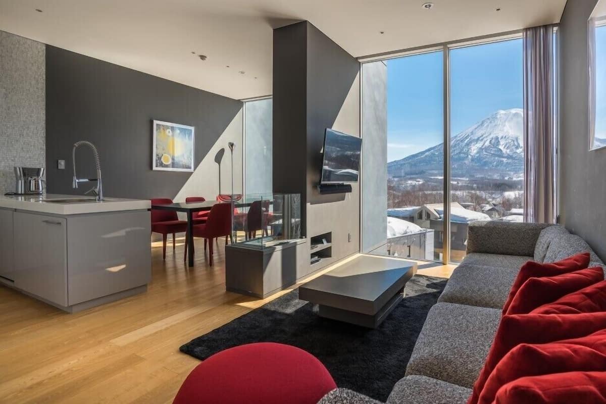 A modern living room with a sofa, red chairs, a dining table, a kitchen area, and a large window with a mountain view.