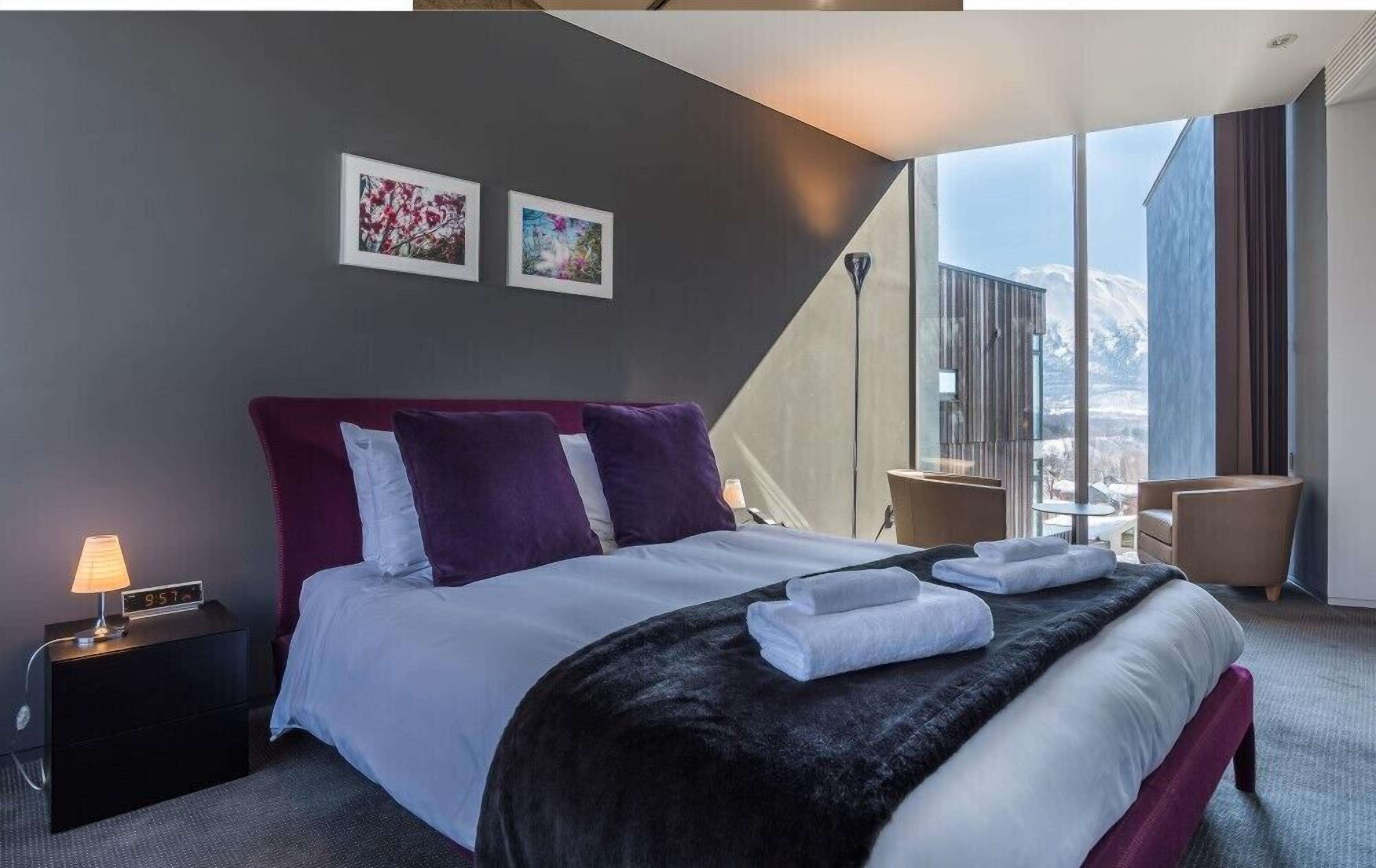 A modern hotel room with a large bed, a nightstand, a lamp, two framed pictures, a chair, and a view of the mountains through large windows.