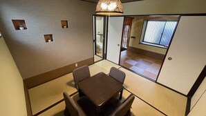 A room with tatami flooring, a traditional Japanese-style table with chairs, a hanging light fixture, and a sliding door leading to another room. - Kanon House (Kyoto)
