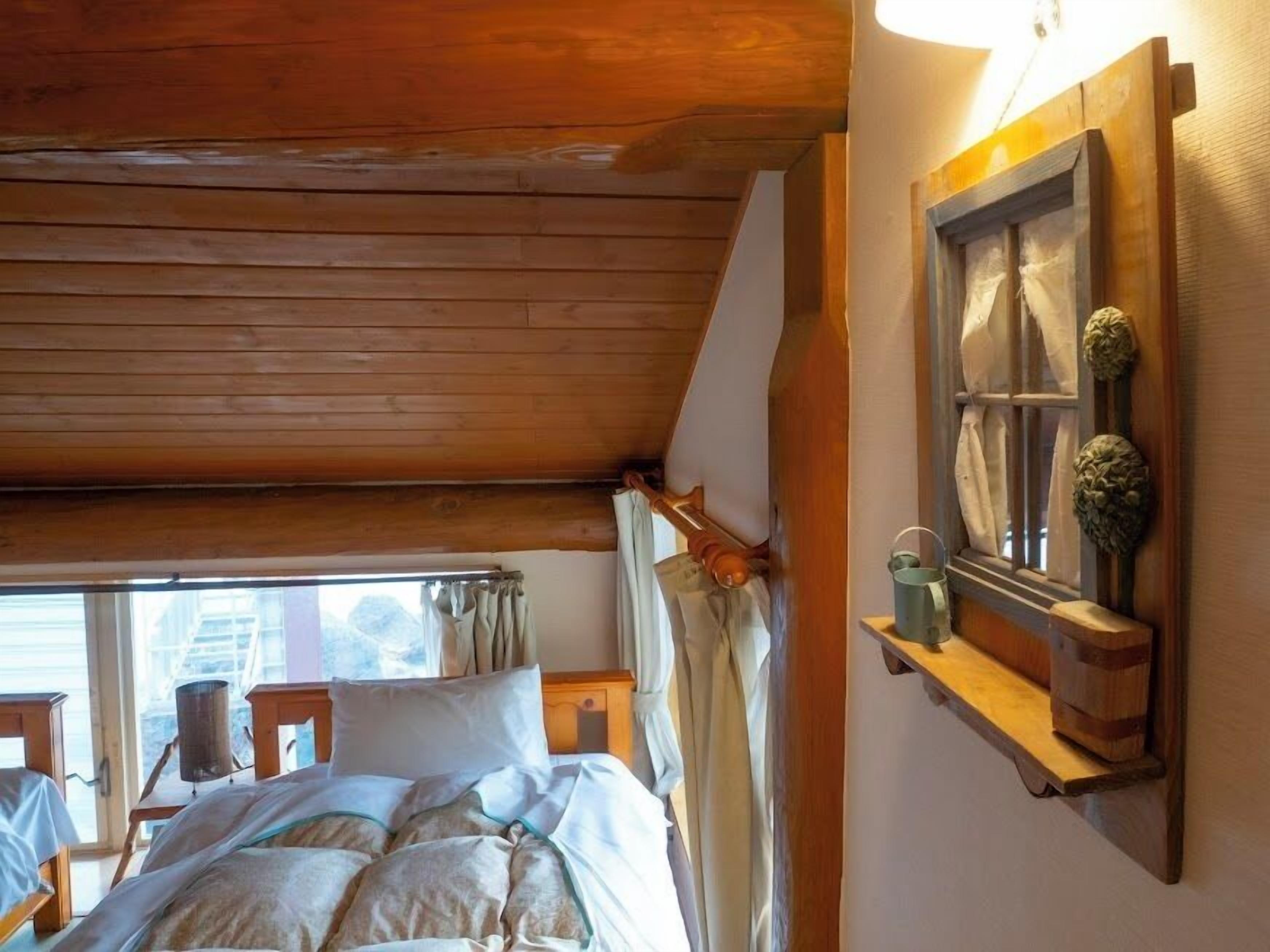 A wooden ceiling, a bed with pillows, a window with curtains, and a shelf with decorative items.