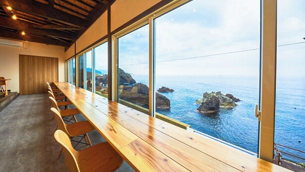 Una larga mesa de madera con sillas en una habitación con ventanas amplias que dan a una costa rocosa.