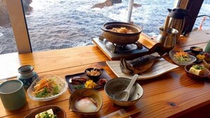 Una mesa de madera con diversos platos, un tazón de sopa y una olla humeante, vista desde una ventana con vistas al mar.