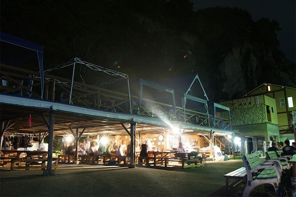 Un espacio de comedor al aire libre con mesas y bancos, una estructura cubierta y un edificio con fachada verde.