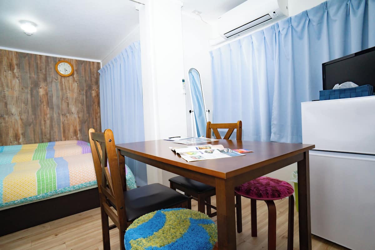 Una habitación con cama, una mesa de madera con sillas, un espejo y un refrigerador pequeño.