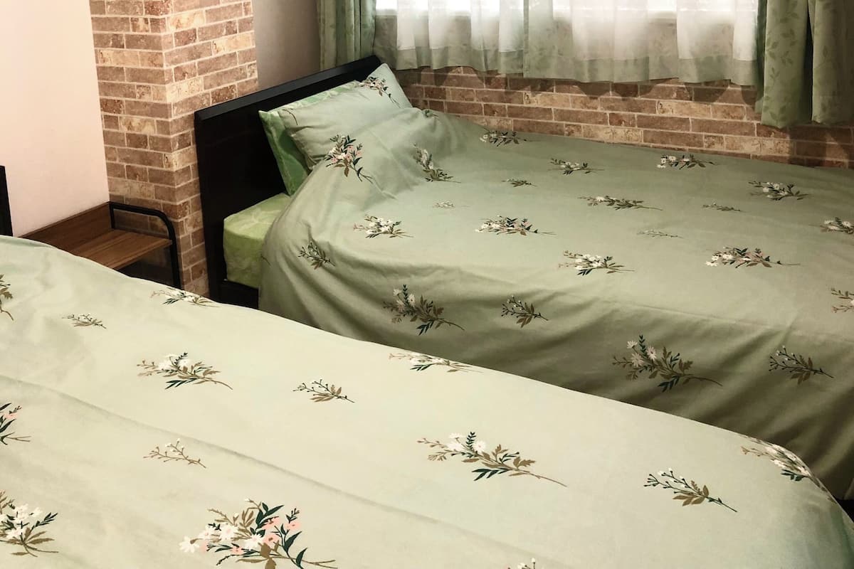 Una habitación con pared de ladrillo, ventana con cortinas verdes y dos camas con ropa de cama estampada.