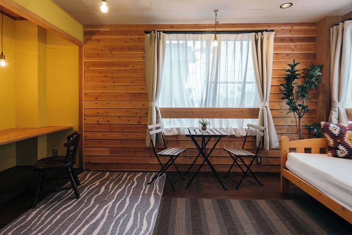 A room with wooden walls, a window with curtains, a small table with chairs, a bed, and a potted plant.