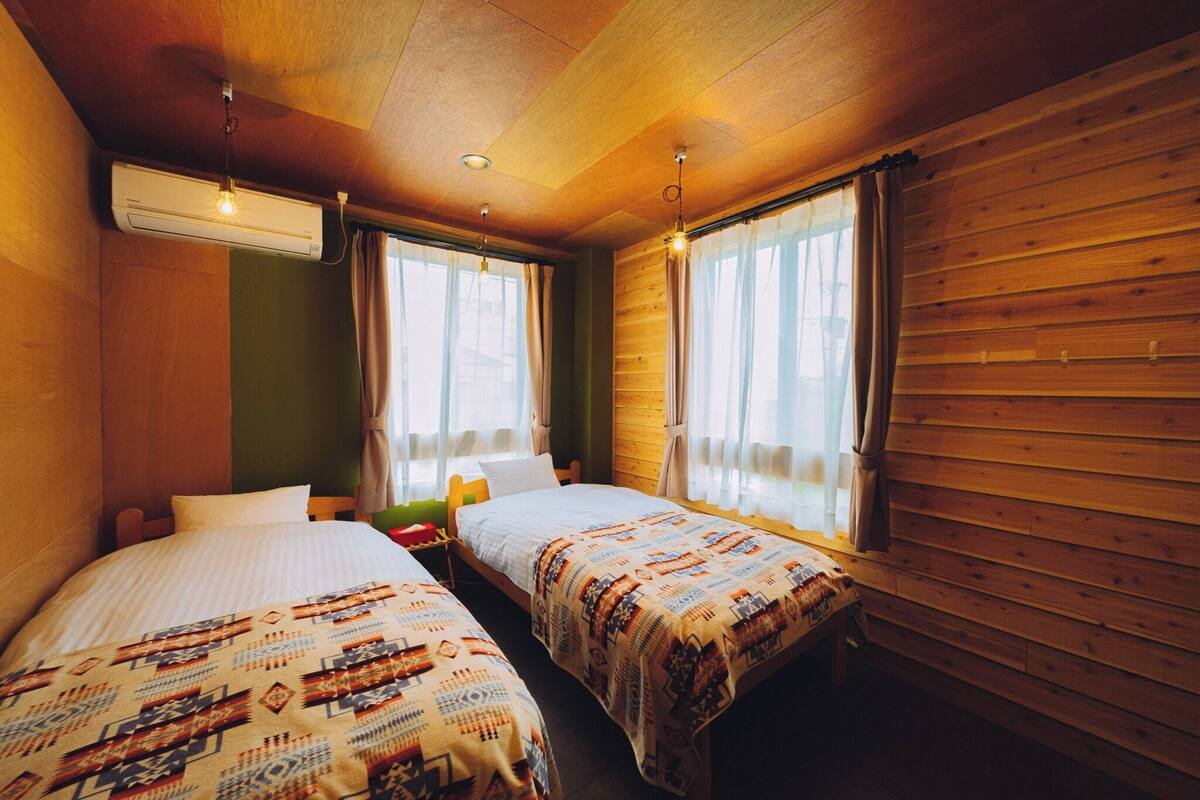 A room with two beds, wooden walls, a window with curtains, an air conditioning unit, and a small table.