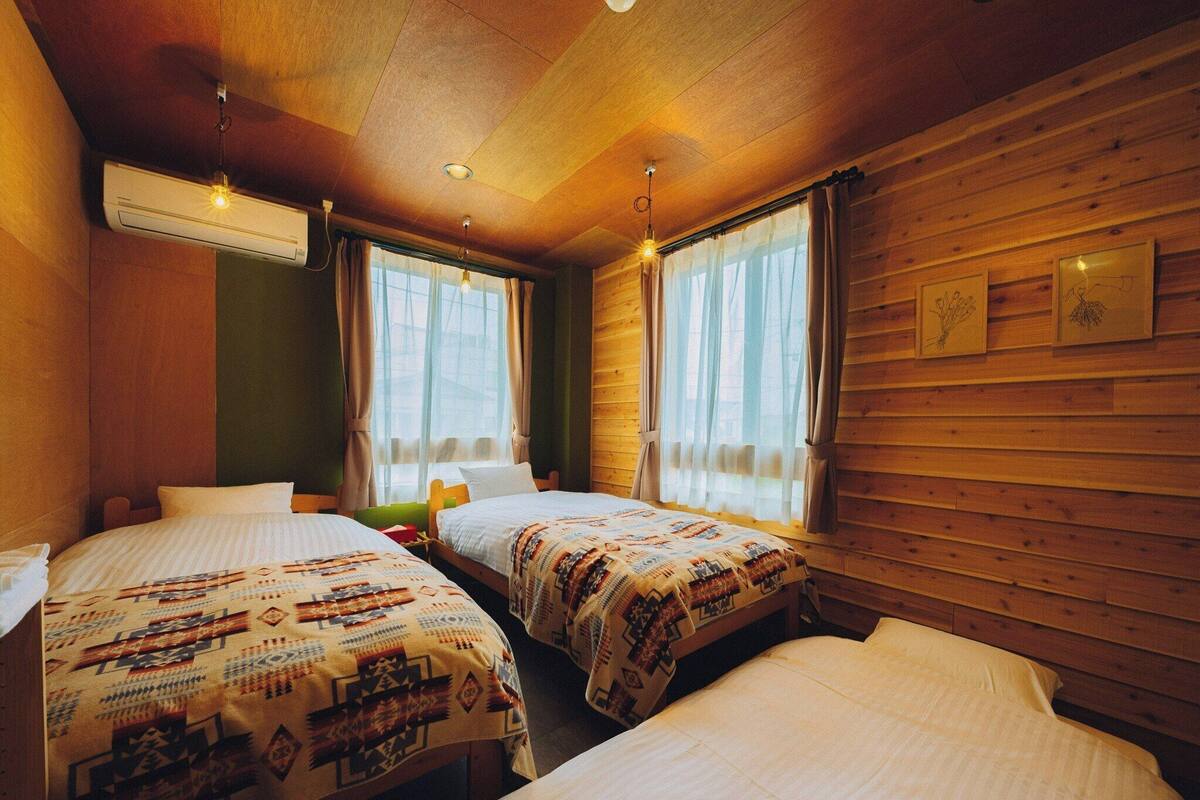 A room with two beds, wooden walls, a ceiling fan, and a window with curtains.