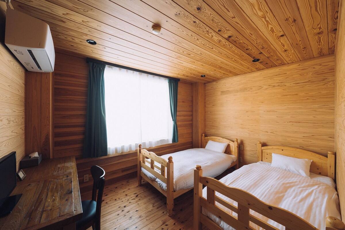A wooden cabin room with two beds, a table, and a window with curtains.