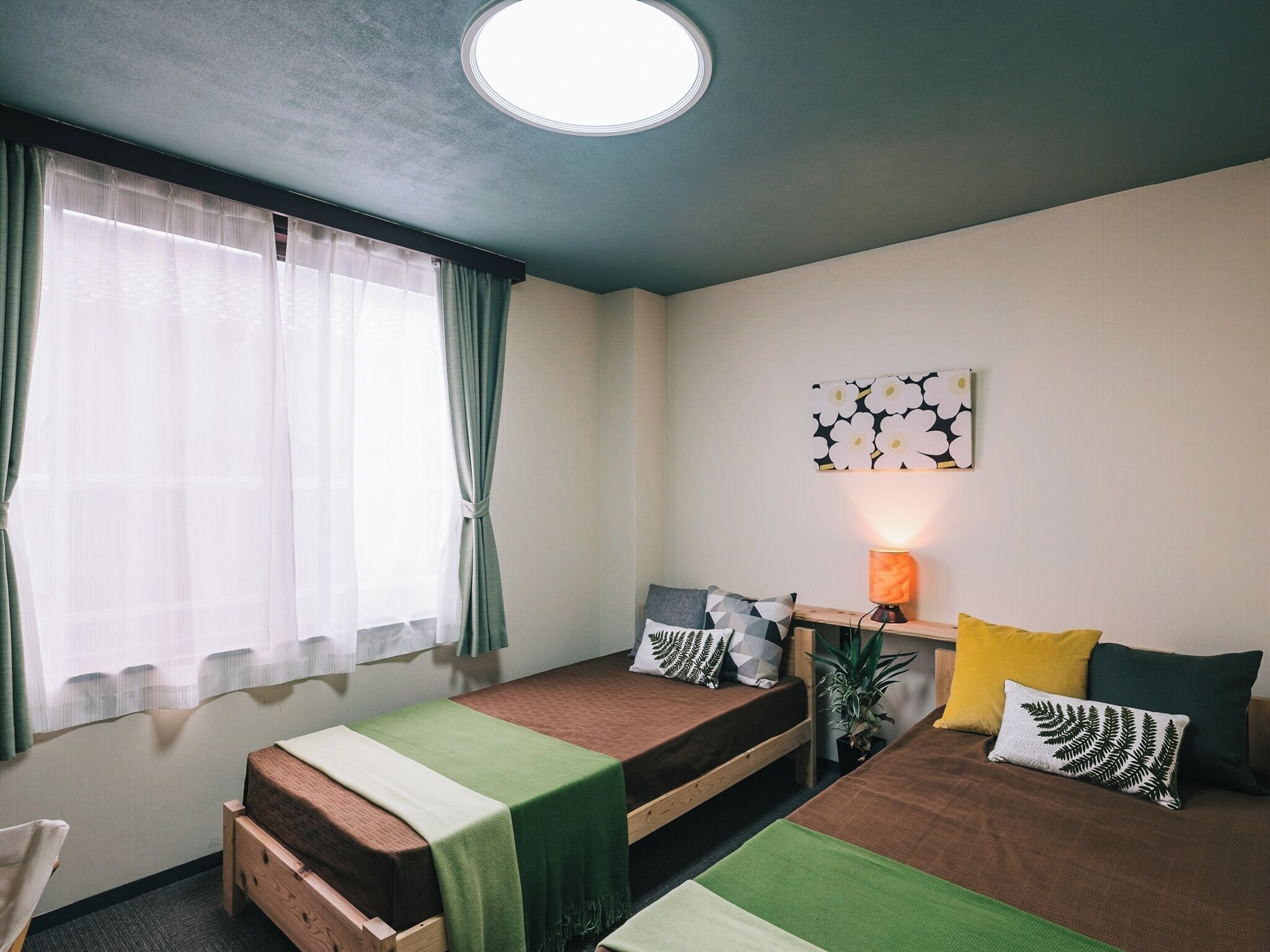 A room with two beds, a desk with a lamp, a window with sheer curtains, and a wall-mounted artwork.