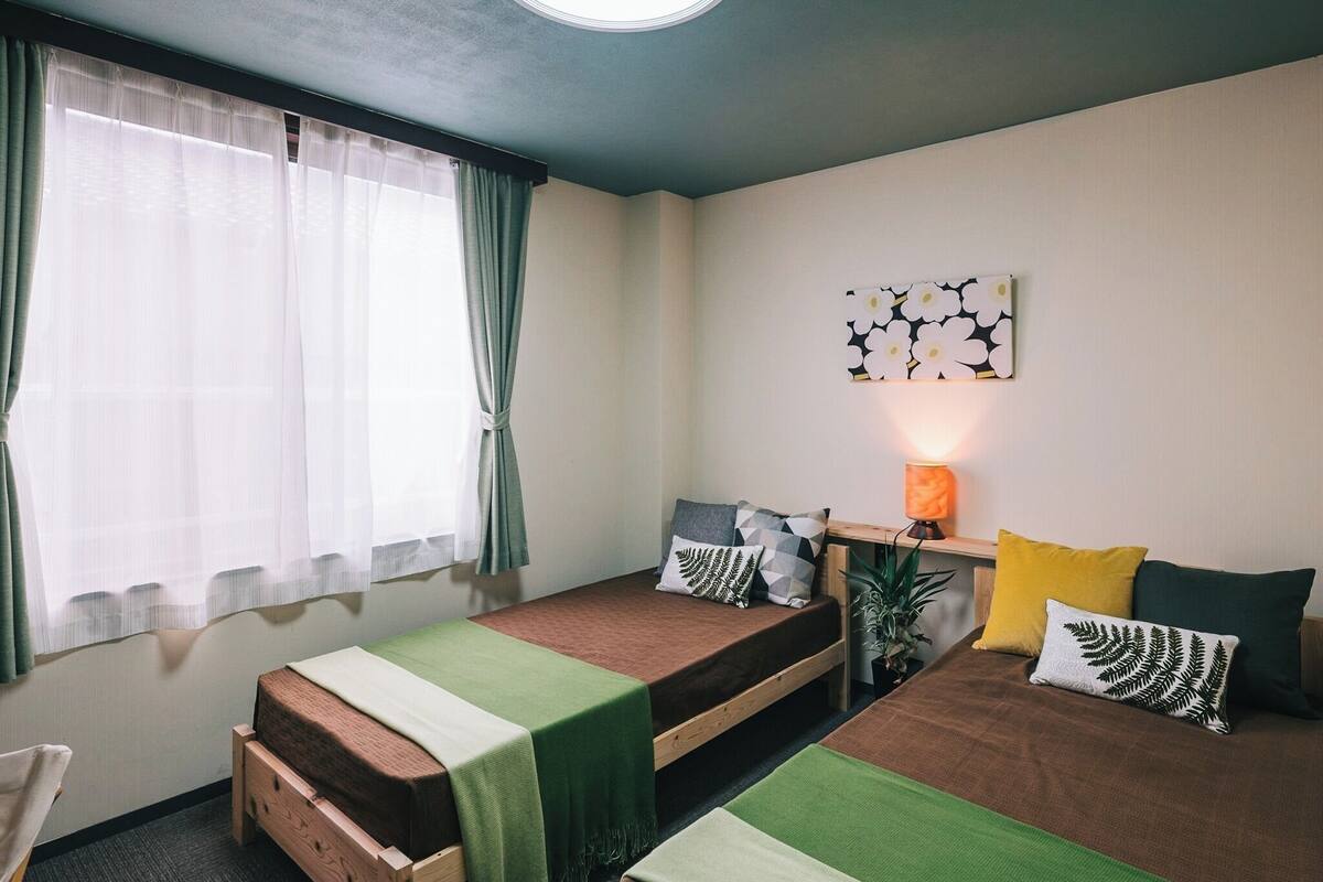 A room with two beds, a desk with a lamp, a window with sheer curtains, and a wall-mounted artwork.