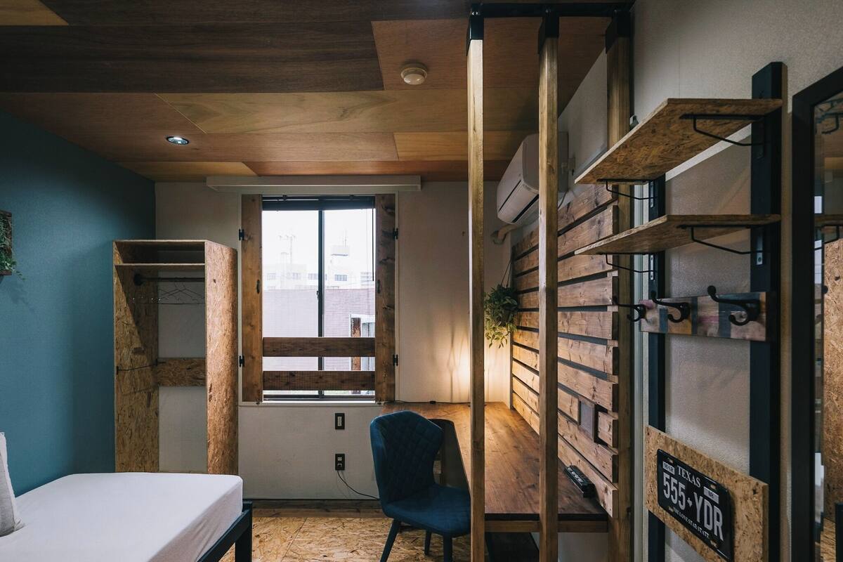 A room with a bed, a chair, a wooden wall unit with hooks, and a window with a view of buildings.