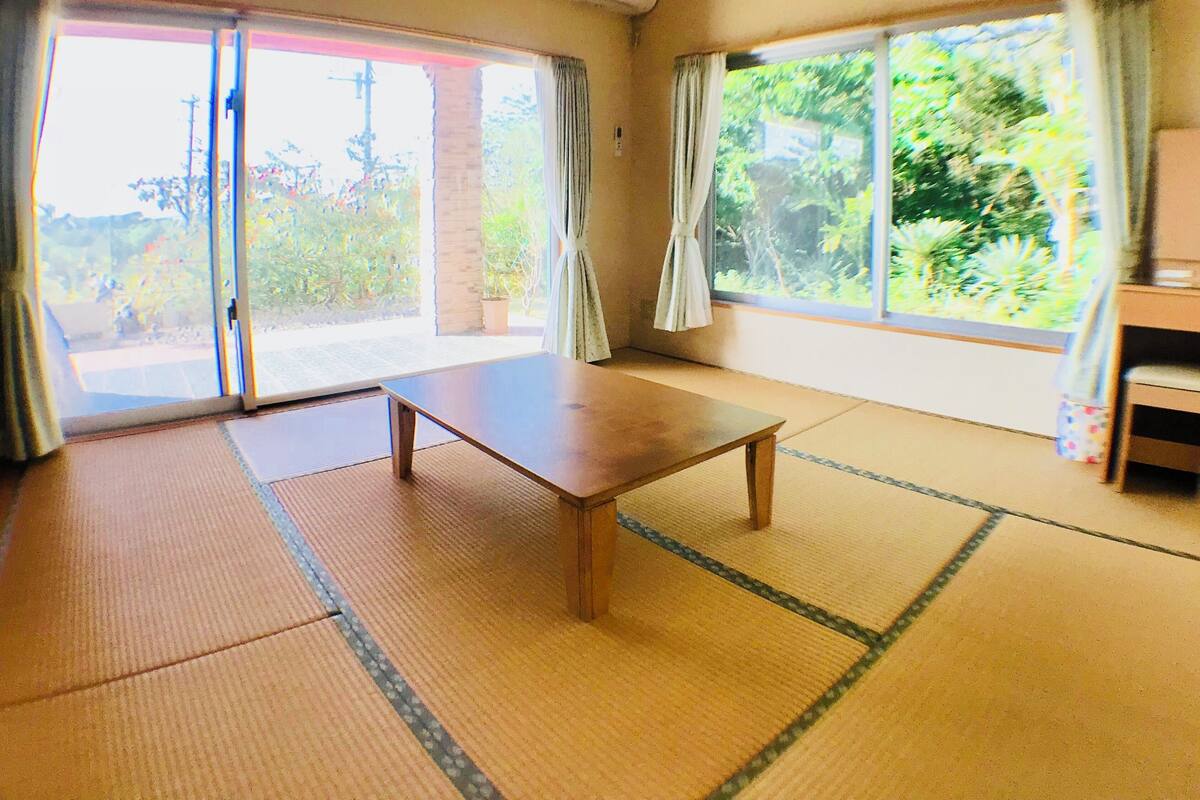 A room with tatami mats, a wooden table, sliding glass doors, and a view of greenery outside.