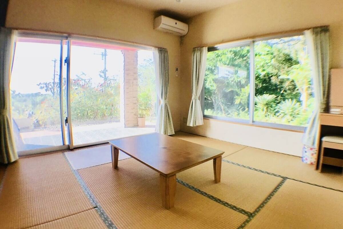A room with tatami flooring, a wooden table, sliding glass doors, and a view of greenery.