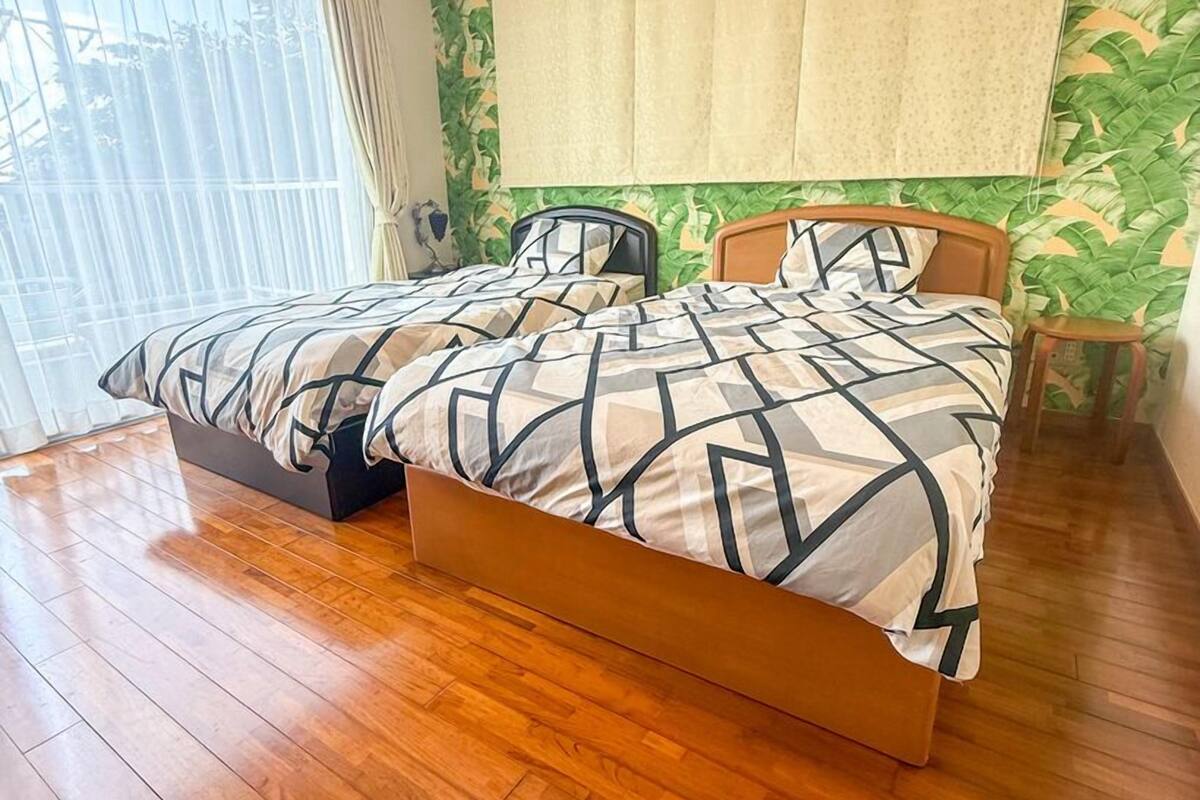 A double bed with a geometric patterned bedspread, a wooden headboard, and a large window with sheer curtains.