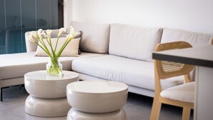 Una sala moderna con un sofá, dos mesitas redondas blancas, un jarrón con flores y una silla de madera.