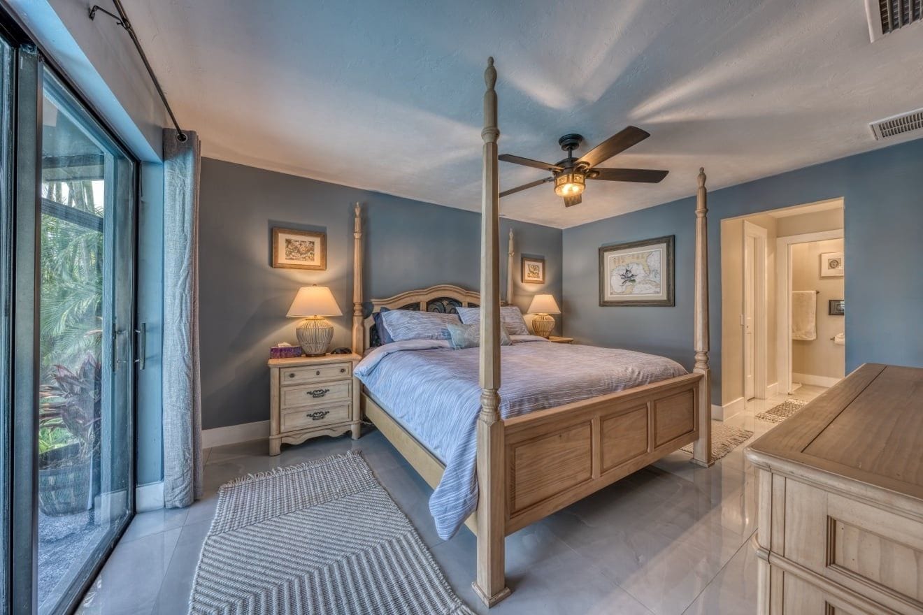 A bedroom with a four-poster bed, bedside lamps, a ceiling fan, and a view through a sliding glass door.