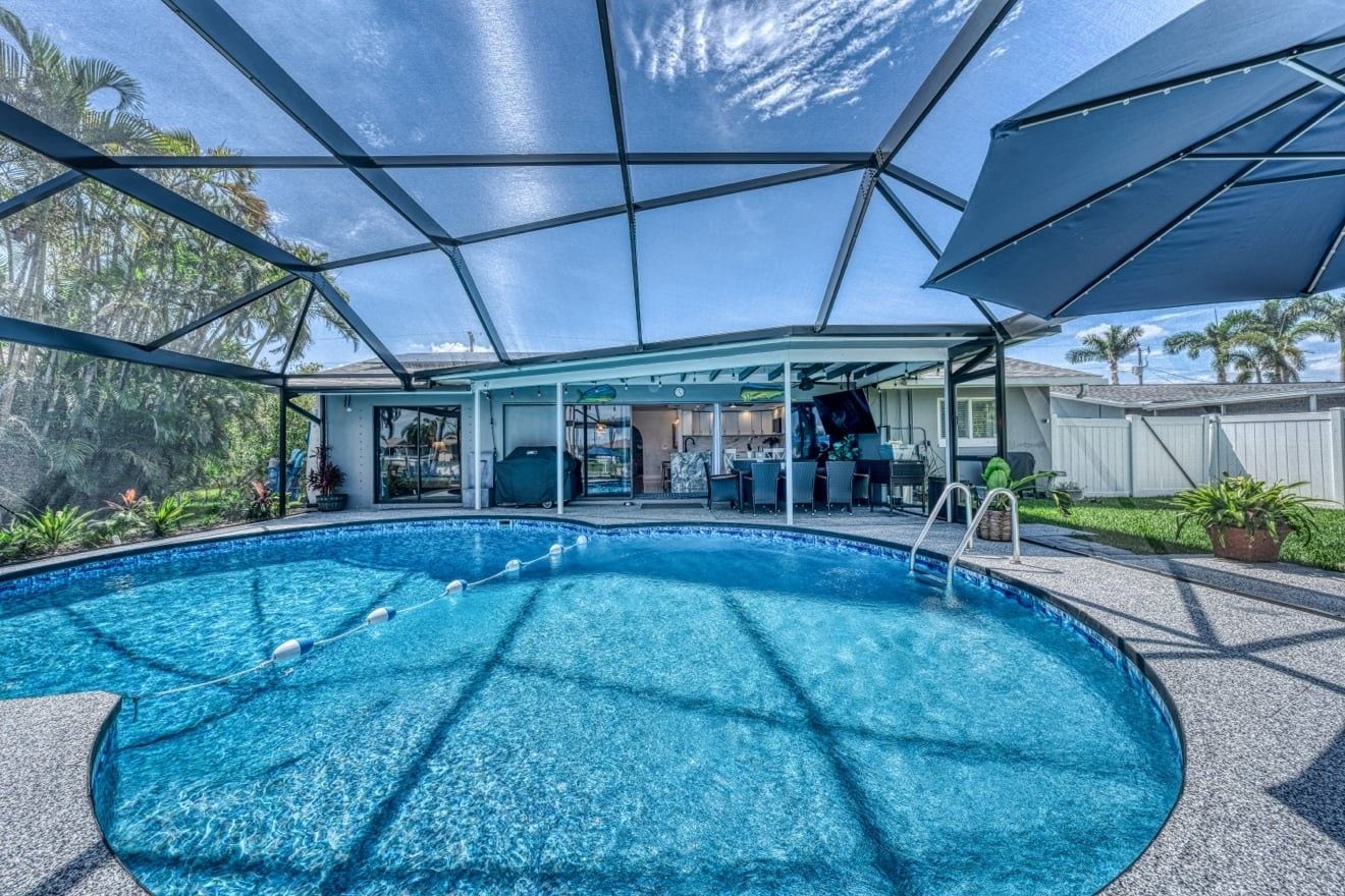 A swimming pool with a covered area, surrounded by a fence, and palm trees in the background.