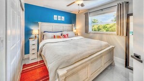 Ein Schlafzimmer mit einem Bett, einem Nachttisch, einer Lampe, einem Fenster mit Jalousien und einem Deckenventilator.