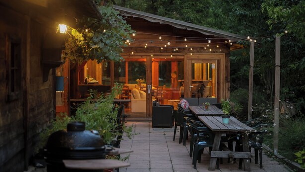 An outdoor patio area with wooden tables and chairs, string lights, and a view of the interior through glass doors. - Hotel les 5 saisons (Freissinières)