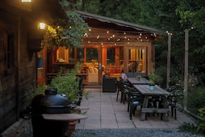 Un área de patio al aire libre con mesas y sillas de madera, luces colgantes y vista al interior a través de puertas de vidrio.