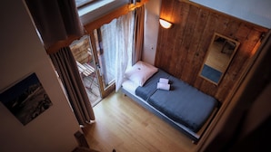 A small, single-bed room with a wooden floor, a window with curtains, a wall-mounted light, and a small mirror. - Hotel les 5 saisons (Freissinières)