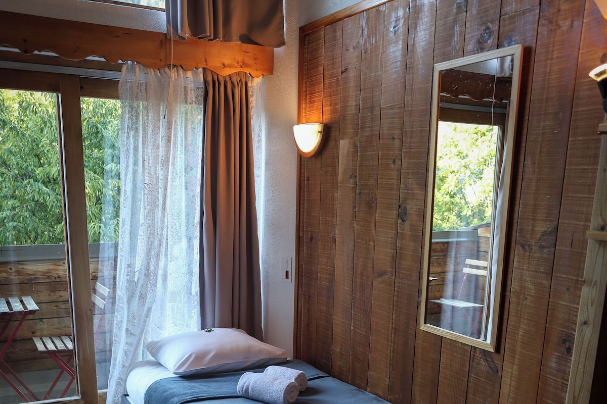 Una cama individual con colchón y almohadas, suelo de madera, ventana con cortinas y una lámpara fijada a la pared.