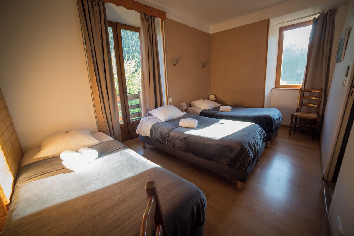 Una habitación de hotel con tres camas, una silla de madera y un ventanal con cortinas.