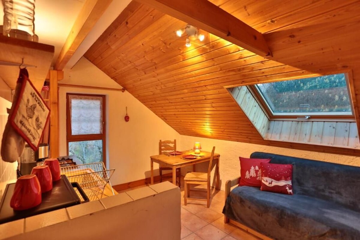 Una habitación acogedora con techo inclinado, una mesa de comedor con sillas, un sofá con cojines y una ventana con vistas a árboles.