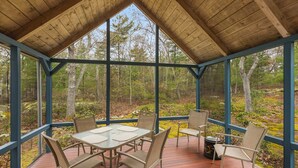Une terrasse extérieure couverte, dotée d’une table et de chaises, et entourée de murs de verre offrant une vue sur une zone boisée.