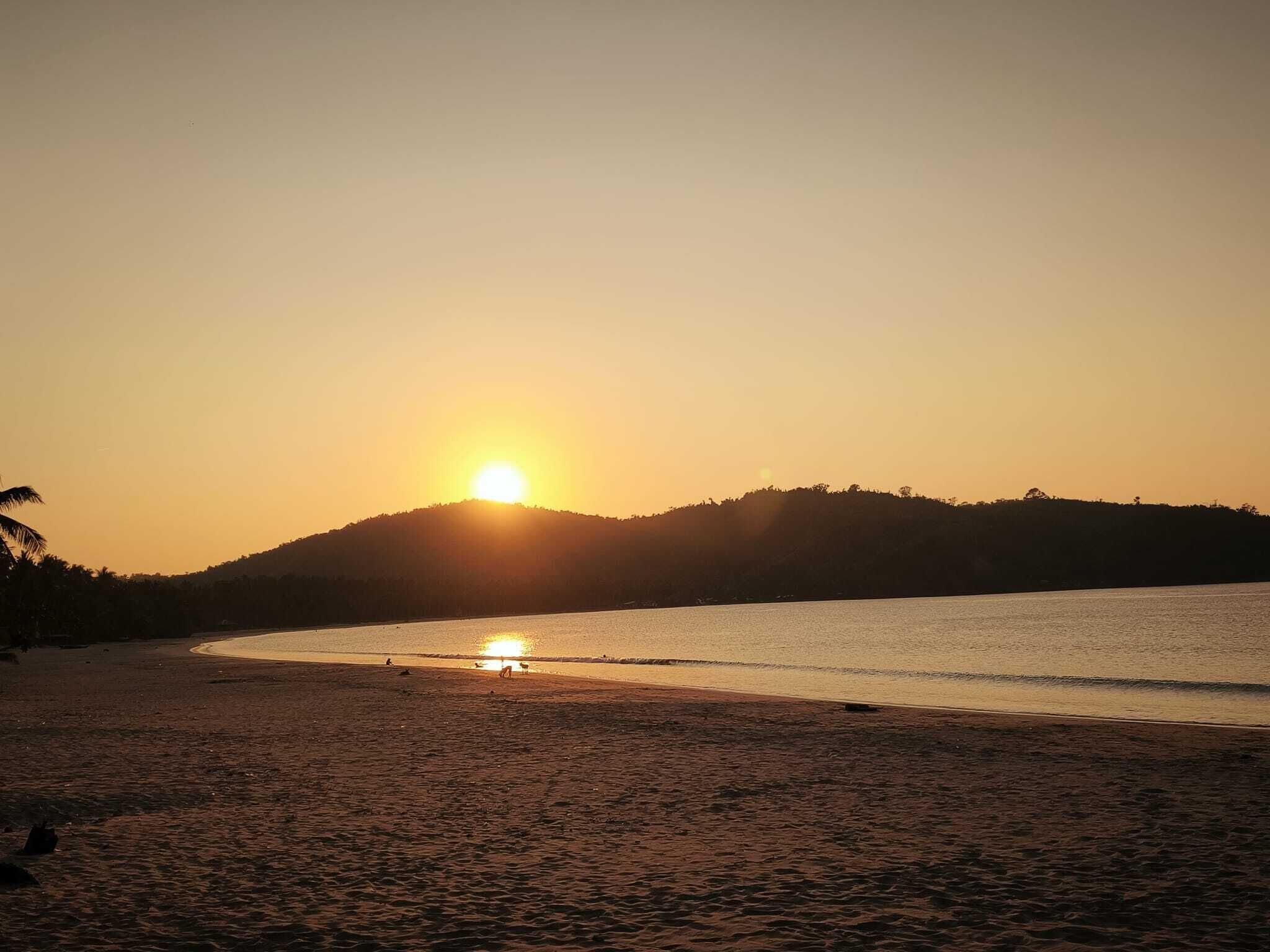 Une plage au coucher du soleil, avec une eau calme et une colline qui se dessine à l’horizon.