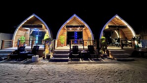 Three wooden dome-shaped structures with outdoor seating and potted plants, situated on a sandy beach at night.
