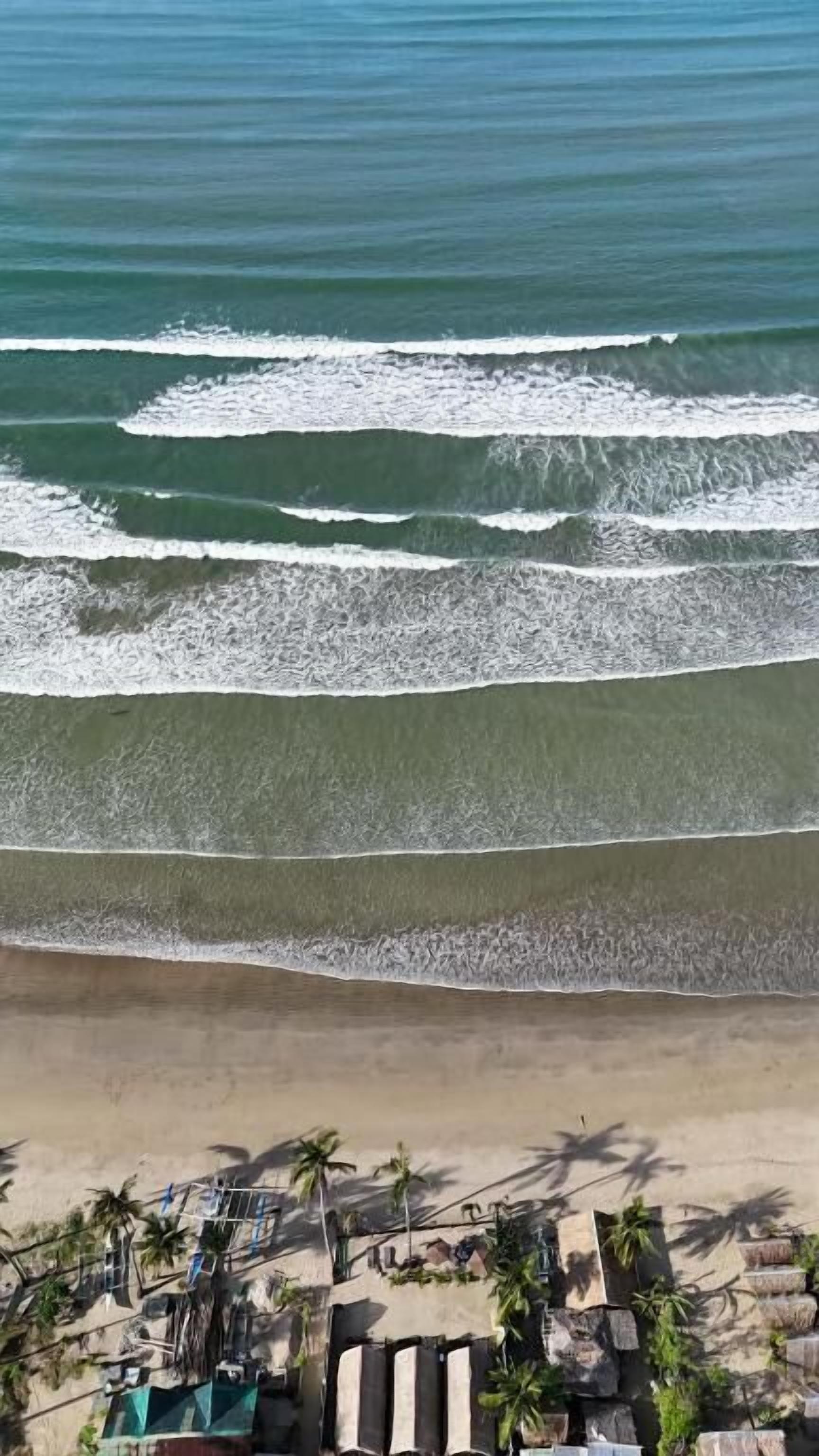 A beach with waves, a sandy shore, and a structure with umbrellas and plants.