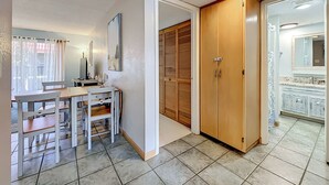Ein Zimmer mit Esstisch, Stühlen, einem Holzschrank, einem Badezimmer mit Spiegel und Waschbecken sowie einem Fenster mit Vorhängen.