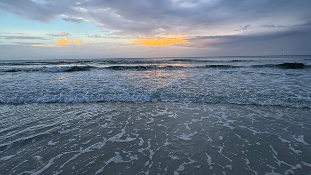 Ein Strand mit Wellen, bewölktem Himmel und Sonnenuntergang.