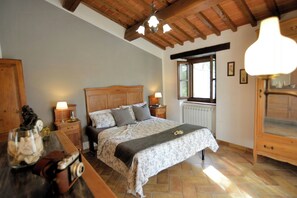 Ein Schlafzimmer mit einem Holz-Kopfteil, einem Bett mit floralem Überwurf, zwei Nachttischlampen, einem Nachttisch aus Holz, einer Kommode und einem Fenster mit Jalousien.