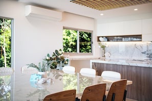A modern dining area with a glass table, wooden chairs, and a view of greenery outside.