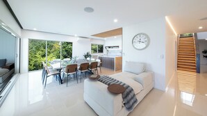 A modern living space with a dining area, kitchen, and a large window offering a view of greenery. - 180 Degree Ocean Views - 5 Bedroom - Heated Magnesium Swimming Pool (Airlie Beach)