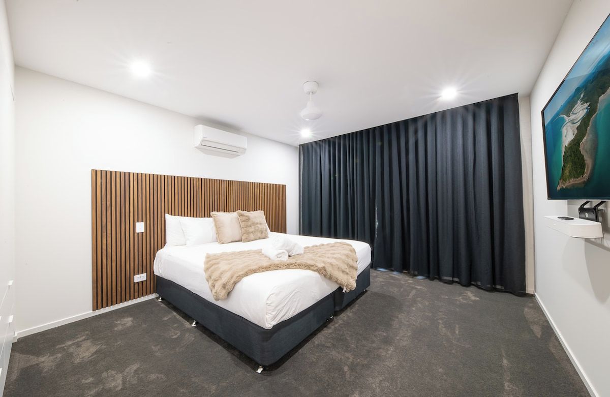 Une chambre d’hôtel moderne avec un grand lit, une tête de lit en bois, un rideau foncé et un tableau au mur.