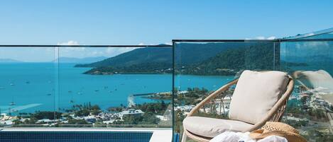 A glass-enclosed balcony with a view of a coastal area, a pool, and a wicker chair with a cushion.