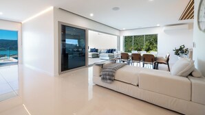 A modern living room with a sofa, chairs, a dining table, and a large window with a view. - 180 Degree Ocean Views - 5 Bedroom - Heated Magnesium Swimming Pool (Airlie Beach)