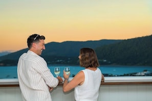 A couple standing by a balcony overlooking a body of water with hills in the background.