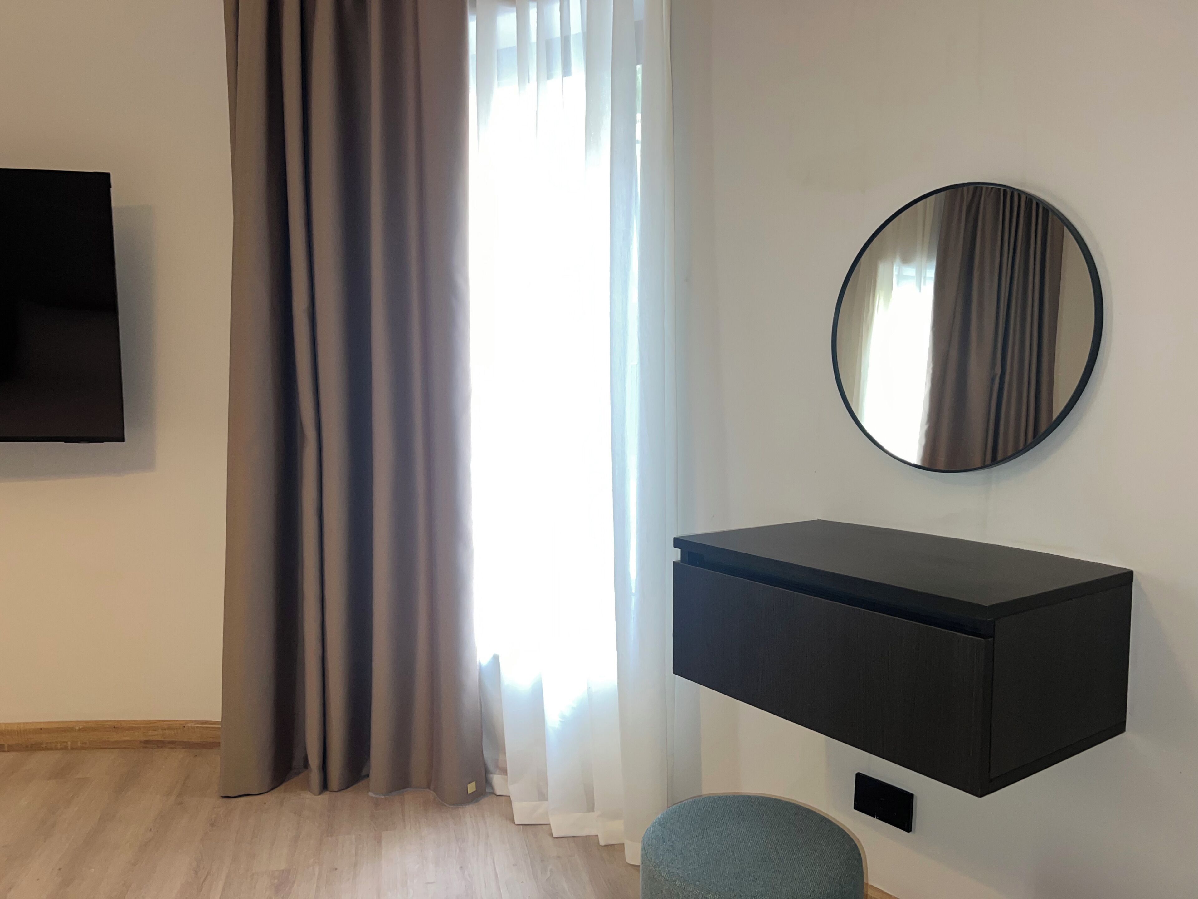 A modern room with a flat-screen TV, a round mirror, a dark console unit, and a window with curtains.
