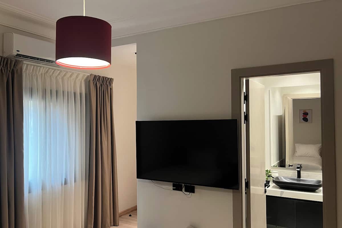 Ein Hotelzimmer mit Wand-Fernseher, einer modernen Pendelleuchte, einem Badezimmer mit Waschbecken und Spiegel sowie einem Fenster mit transparenten Vorhängen.