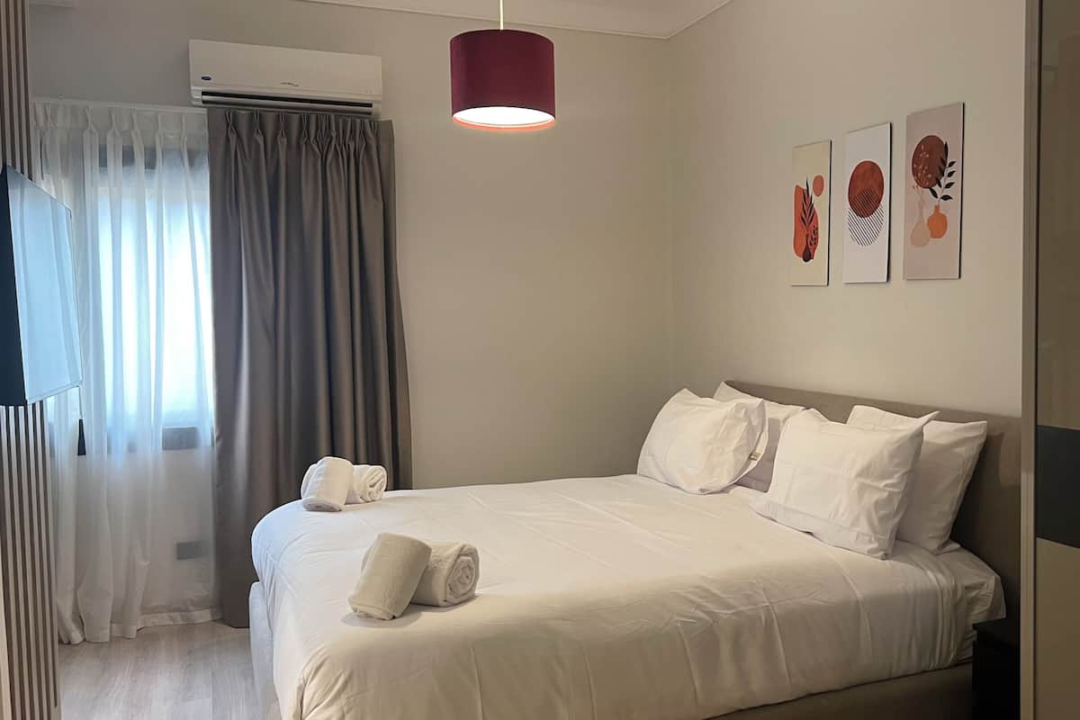 Ein Hotelzimmer mit einem Bett, einer Wandklimaanlage, einer Deckenleuchte und drei gerahmten Kunstwerken an der Wand.