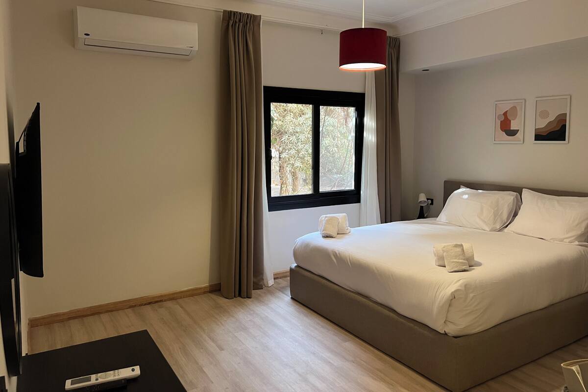 Ein Hotelzimmer mit Bett, Fernseher, Klimaanlage, einem Fenster mit Vorhängen und einer roten Pendelleuchte.